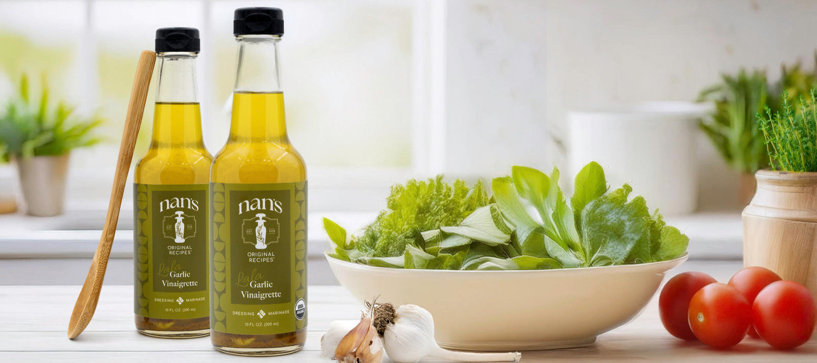 Two bottles of Lala Vinaigrette displayed on a kitchen counter surrounded by fresh produce, including leafy greens, tomatoes, and garlic.